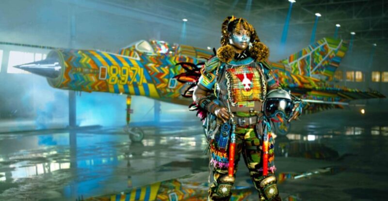 A MiG-21 military flight suit is being modeled by a woman of African descent. She is covered in a brightly colored tribal-inspired armor featuring colorful beads.